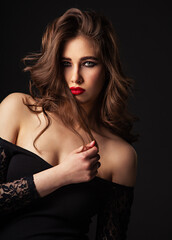 Fototapeta premium Beautiful bright makeup woman with long brown curly volume brown hair style, red lipstick, and black smokey eyes looking vamp and passion on black background. Closeup