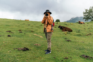 Caucasian woman in a brown coat and hat taking a picture of the camera in a field with cows in the...