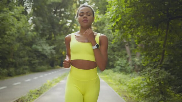 Young sporty african american woman in bright sportsuit running in morning, looking at camera, jogging in public park