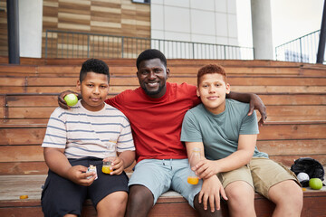Portrait of positive African-American father sitting on wooden bleacher bench and embracing sons at sports match