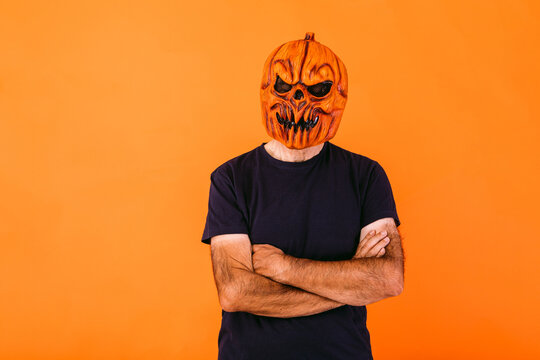 Man wearing scary pumpkin latex mask with blue t-shirt with crossed arms, on orange background. Halloween and days of the dead concept.