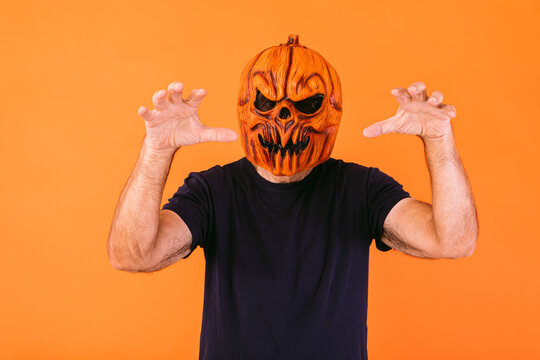 Man wearing scary pumpkin latex mask with blue t-shirt scares with his hands, on orange background. Halloween and days of the dead concept.