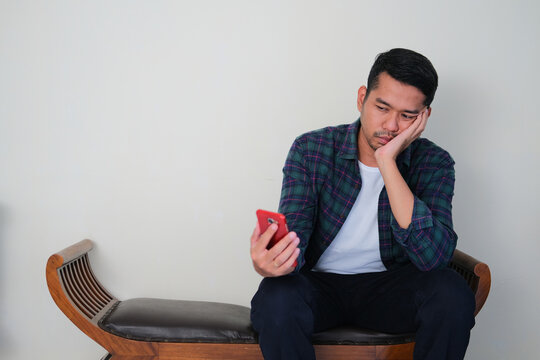 Adult Asian man sitting in a chair and showing boring gesture while looking to his mobile phone