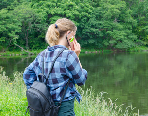 Rear view of a conversation on a mobile phone in the open air in a park by the lake. The girl is holding a mobile phone in her hands, using a digital device, looking into the distance. selective focus