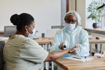 Medical nurse in PPE suit checking body temperature of young woman with non-contact infrared...