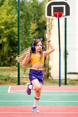 Schoolgirl in wireless headphones listening to music wearing fashionable sportswear jumping on sports ground. Rehearsal for a cheerleading performance