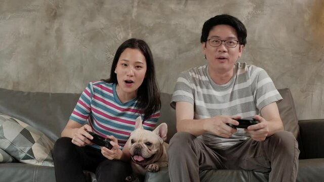 Asian couple is playing video games console, excitedly and having fun on sofa at home, control joystick to competition, pet dog (French Bulldog) nearby. It's happiness in family lifestyle.