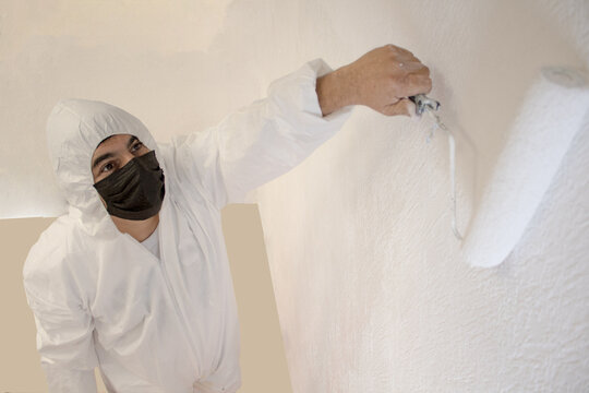 Hombre j&oacute;ven trabajador pinta una pared blanca con un traje de protecci&oacute;n blanco y cubrebocas KN95 negro