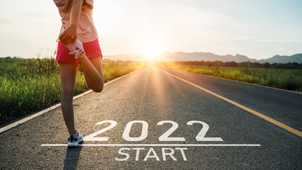 New year 2022 or start straight concept.word 2022 written on the asphalt road and athlete woman runner stretching leg preparing for new year at sunset.Concept of challenge or career path and change. - obrazy, fototapety, plakaty