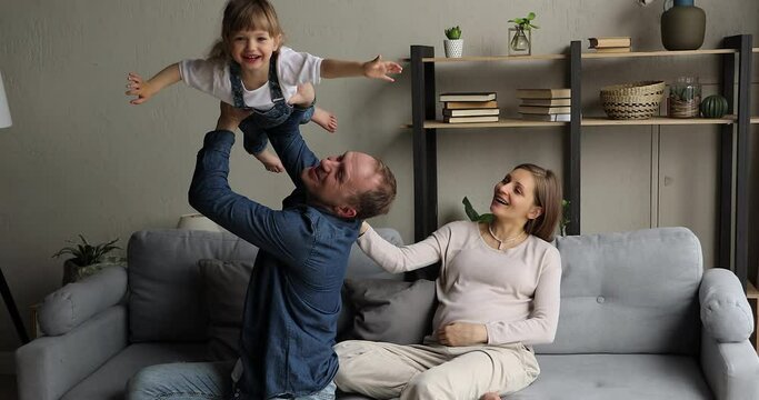 Pregnant woman sit on sofa with husband play with little 4s cute daughter lifts her on arms, cheerful kid girl imagine like fly in air. Happy family enjoy playtime at home. Leisure, parenthood concept