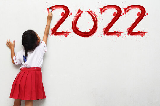 Little girl holding a paint brush painting happy new year 2022 on a white wall background, Back to school idea concept