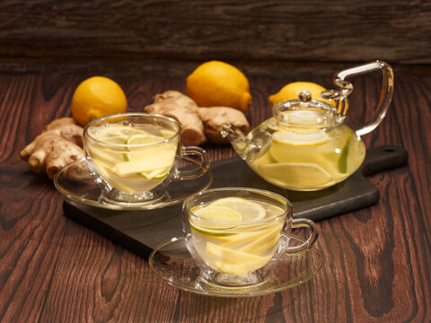 Ginger and lemon tea in glass cups and teapot on a burned wood cutting board over brown rustic wood background