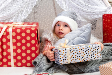 portrait of a cute little girl child wearing a silver color Christmas hat lying between gift boxes