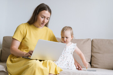 Curious blond blue-eyed baby girl looking at mom laptop display while sitting on sofa in living...