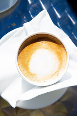 Top view to a hot fresh cup of capuccino in bright sunlight. White mug of hot coffee with milk on white napkin. Traditional Italian breakfast beverage. Vacation in Europe drink. Close up 
