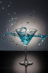 Ice-cube falling into a splashing blue cocktail martini glass.