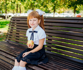 Back to school. A cute little schoolgirl is sitting on a bench in the school yard and holding a green apple.