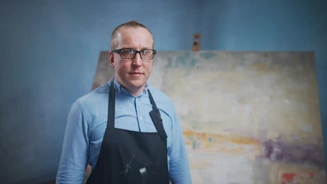 Slow motion, portrait of an adult male artist standing against the background of a large canvas, a man stands in his art studio and looks at the camera, creative person in a blue shirt and glasses at