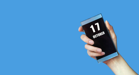 october 17. 17th day of the month, calendar date.Woman's hand holds mobile phone with blank screen on blue isolated background.Autumn month, day of the year concept