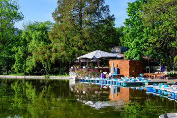 Fototapeta na wymiar Vivid green landscape with old large linden trees and small boats near the lake in Cismigiu Garden (Gradina Cismigiu), a public park in the city center of Bucharest, Romania, in a sunny spring day.