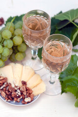 Two glasses of white wine, bunches of grapes, a young vine, cheese and walnuts on a light wooden background. a vertical snapshot.