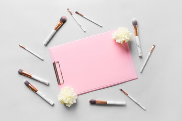 Clipboard with blank sheet of paper, makeup brushes and flowers on light background