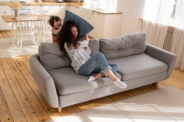 Fototapeta premium Happy mom and cheerful child playing in living room overjoyed pillow fight throwing cushions sitting on sofa at home. Loving parent and preschool kid enjoy time together. Funny family activity concept