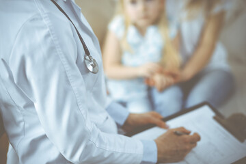 Fototapeta premium Doctor and patient. Pediatrician using clipboard while examining little girl with her mother at home. Sick and unhappy child at medical exam. Medicine concept