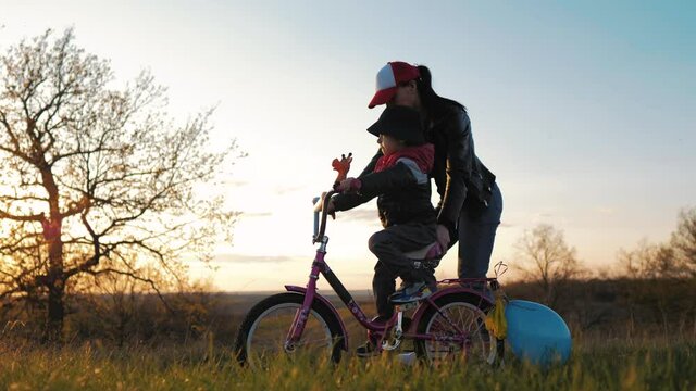 Childhood bike concept. Mother teaching son to ride bicycle. Happy cute boy learn to riding a bike in park at sunset time. Young mom teaching son to ride bike first time on countryside rural road.