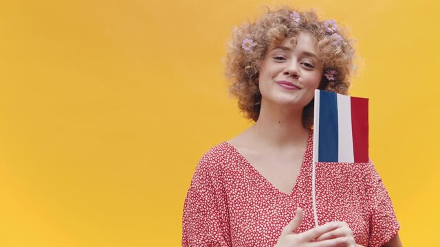 Attractive girl holding a France Flag in her hand. Girl in a joyful mood over yellow background. Dressed in a casual pink dress and wearing flowers in her hair. Learn French Concept.
