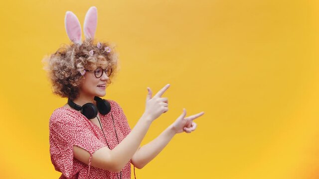 Attractive young girl wearing cute bunny ears and spectacles in a joyful mood. Headphones around her neck. Girl smiling and pointing at something. Bright yellow background. Studio shot.