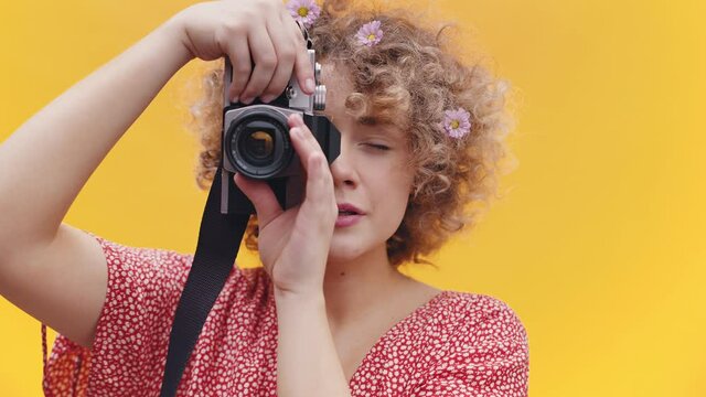 Attractive girl holding a vintage camera taking a picture. Girl in a joyful mood over bright yellow background studio. Dressed in a casual pink shirt wearing flowers in her hair. Photography concept.