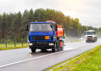 Fototapeta na wymiar Two tanker trucks for transporting petroleum products drive on the highway with their headlights on in cloudy weather and rain in summer. Gasoline transportation concept, dangerous cargo