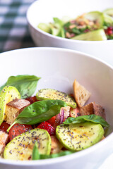 Green salad with cucumber, zucchini and strawberries in a white bowl over green plaid tablecloth.