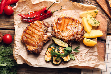 Board with tasty pork steaks and grilled vegetables on wooden background
