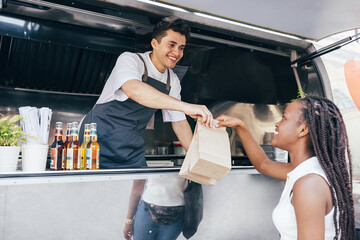 Smiling salesman giving packages with street food to a female customer. Food truck owner serves the...