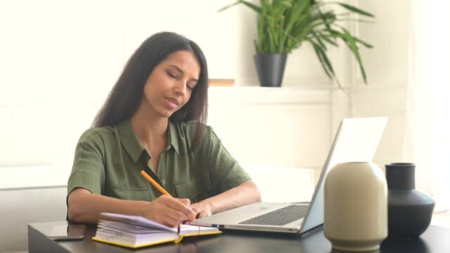 Freelance woman using laptop for remote work from home. Indian female student in casual wear sits at the desk, watching online webinars or training and taking notes, writes down