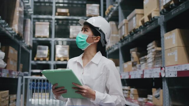 Asian woman warehouse worker wearing hardhat face mask Active checking products in factory. Businesswoman store shelf information on clipboard. wearing mask to prevent COVID19 and dust in factory