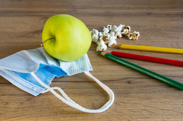 a green apple near white popcorn lies on a protective mask near colored pencils on a wooden table