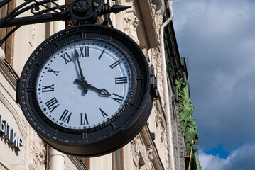 Fototapeta na wymiar An old black round clock hangs against the background of a city house, sculptures and a dark sky