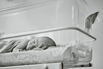 Fototapeta na wymiar A newborn baby with a maternity hospital bracelet on his arm is sleeping in a crib. A newly born child in a clinic bed behind a transparent glass