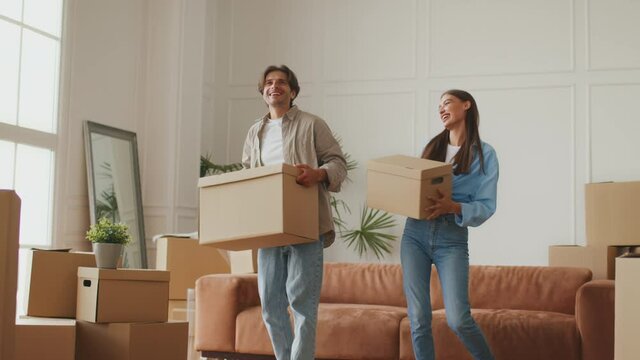 Joyful young married couple celebrating moving to their house, dancing with cardboard boxes in own apartment