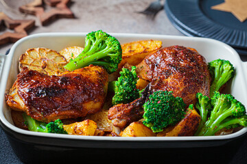 Baked chicken legs with potatoes, broccoli and garlic in baking dish, dark background. Healthy festive food concept.