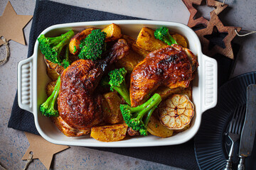 Baked chicken legs with potatoes, broccoli and garlic in baking dish, dark background. Healthy festive food concept.
