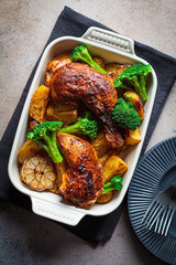 Baked chicken legs with potatoes, broccoli and garlic in baking dish, dark background. Healthy festive food concept.