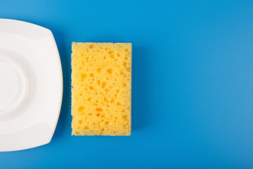 Dishwashing minimal creative concept. Close up of white shiny clean plate and yellow cleaning sponge against blue background with copy space. 