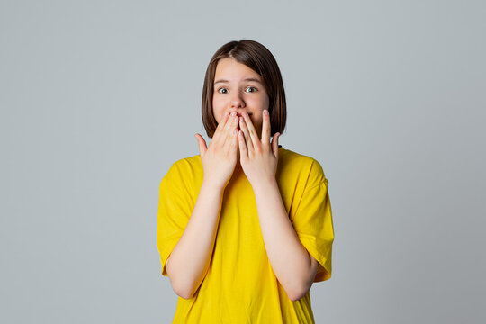 Portrait of a scared teen girl in yellow casual t shirt, looking at camera with wide opened eyes, covering mouth her hands, stand over light grey background