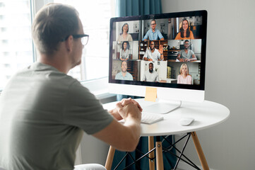 Morning meeting on the distance. Back view over shoulder of guy to the computer screen with a lot of diverse people on it, work team brainstorming, discussing together, virtual meeting colleagues