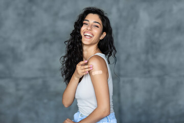 Fototapeta premium Lovely young woman getting vaccinated against covid-19, showing arm with band aid on grey studio background