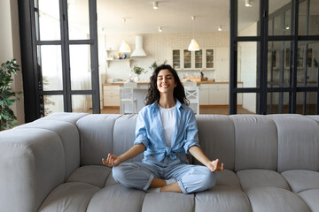 Stress management. Peaceful young lady in casual wear meditating with closed eyes on couch at home